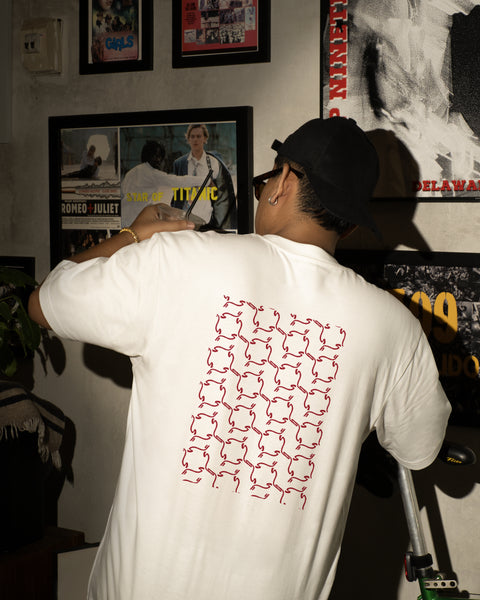 Person wearing a white t-shirt with a red geometric pattern, standing in a room with framed pictures on the wall.
